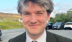 A person with short, tousled dark and grey hair, wearing a suit and tie, stands outdoors near a road with parked cars and grassy hills in the background.