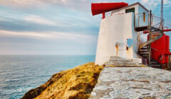 A white lighthouse with a large red foghorn sits on a rocky cliff overlooking the sea under a partly cloudy sky.
