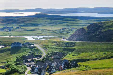 A small cluster of houses sits on a green, hilly landscape with a winding road and water in the background under a cloudy sky.