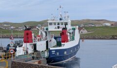 A blue and white ferry docked at a pier, with countryside and houses in the background under a cloudy sky.