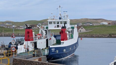 A blue and white ferry docked at a pier, with countryside and houses in the background under a cloudy sky.