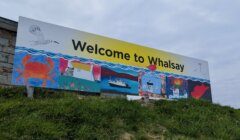 Large "Welcome to Whalsay" sign with colourful illustrations of local wildlife, buildings, and a boat, placed on a grassy hillside under a cloudy sky.