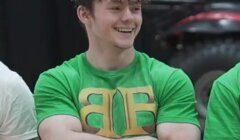 A young man with brown hair, wearing a green T-shirt with yellow lettering, stands with arms crossed and smiles.