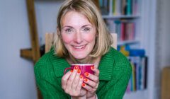 A woman with blonde hair wearing a green jumper holds a red mug and smiles at the camera. Bookshelves are visible in the background.