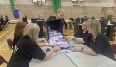 A group of people in a gymnasium sort ballot papers being poured from a box onto a table during an election count.