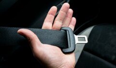A hand holding an unfastened black seat belt inside a car.