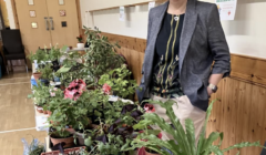 A woman stands indoors beside a long table displaying potted plants and herbs, with signs and wooden panelling in the background.