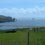 Coastal view in Scotland featuring grassy fields in the foreground, a small inlet, and dramatic sea stacks in the distance under a blue sky.