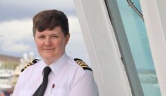 A person in a ship captain’s uniform stands by a white structure, facing the camera with a neutral expression. Some boats are visible in the blurred background.