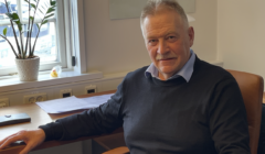 An older man with grey hair and a moustache sits at a wooden desk by a window, wearing a dark jumper over a collared shirt. Papers and a potted plant are on the desk.