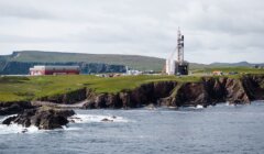 A rocket launch pad stands near the edge of green cliffs by the sea, with buildings and grassy fields in the background under a cloudy sky.