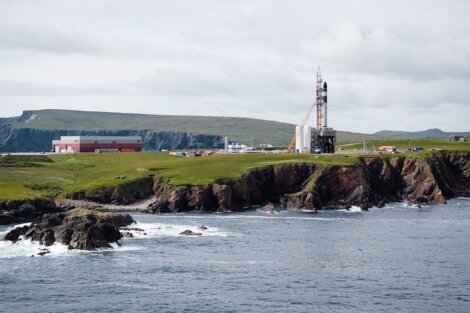 A rocket launch pad stands near the edge of green cliffs by the sea, with buildings and grassy fields in the background under a cloudy sky.