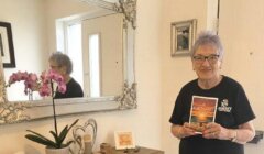 A woman stands in a corridor holding a copy of the book “A Shetland Girl,” with a potted orchid and mirror on the console table behind her.