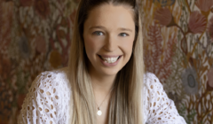 A smiling woman with long, straight blonde hair and a white eyelet top and pendant necklace, posed in front of a floral patterned backdrop.