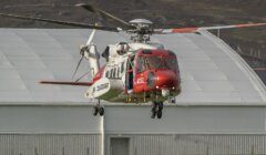 Red and white HM Coastguard helicopter G-MCGL hovering low over grass in front of a large white hangar, with brown hills behind.