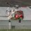 Red and white HM Coastguard helicopter G-MCGL hovering low over grass in front of a large white hangar, with brown hills behind.