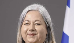 Smiling woman with shoulder-length grey hair in a dark blazer and mustard-yellow scarf, standing in front of a grey backdrop with a blue