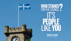 A stone clock tower with crenellated top under a blue sky, with a Shetland (St Andrew's) flag flying from a pole. Text reads "Who stands