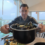 A smiling man in a striped navy jacket holds a large black pot of steamed mussels toward the camera in a bright café with large windows. A