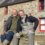 Two smiling men sit side by side on a stone wall in front of a stone house with red-framed windows. The man on the right wears grey waders