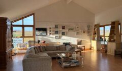 A bright, vaulted-ceiling gallery room with a grey sectional sofa, glass coffee table, framed paintings on a white wall, and large windows