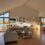 A bright, vaulted-ceiling gallery room with a grey sectional sofa, glass coffee table, framed paintings on a white wall, and large windows