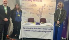 Three people stand beside a "Unlocking Shetland" display board and draped table at an event, with two chairs behind it and a Shetland