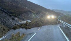 A wet, dim road curves through hills, partly blocked by a landslide of mud and vegetation, with water flowing across the tarmac. Two