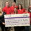 Eight people in red Relay for Life shirts hold a giant cheque for £110,394.13 to Cancer Research UK in front of patterned curtains.