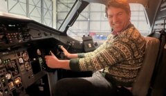 A smiling man in a patterned knitted sweater sits in an aircraft cockpit, one hand on the control yoke, with instrument panels and a hangar
