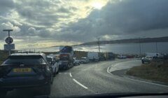 A long line of cars and vans queues along a coastal road beside a calm sea under a cloudy sky, with a "Give way to oncoming vehicles" sign