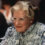 An older woman with curled silver-white hair, clear-framed glasses, pearl earrings and a black-and-white floral blouse sits at a table
