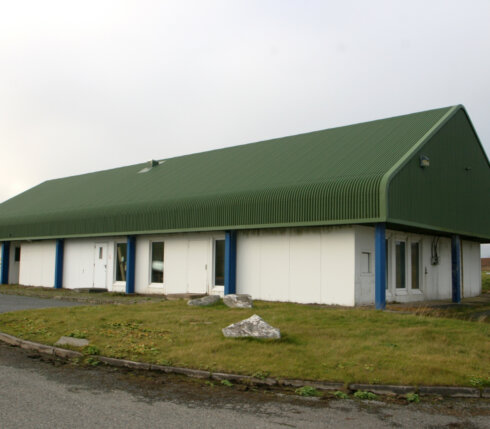 Green-roofed industrial building with multiple doors and an asphalt driveway, under an overcast sky.