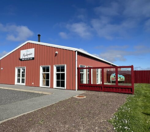 A red and white building labeled "Mackenzies farm shop and cafe" with several glass doors and a fenced outdoor area on a clear, sunny day.