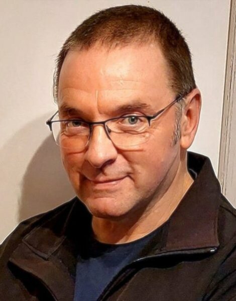 A man with short hair and glasses is wearing a dark jacket and looking at the camera.