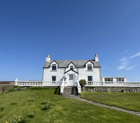 A white, two-story house with a stone walkway, green lawn, and clear blue sky in the background. A white fence borders the front yard.