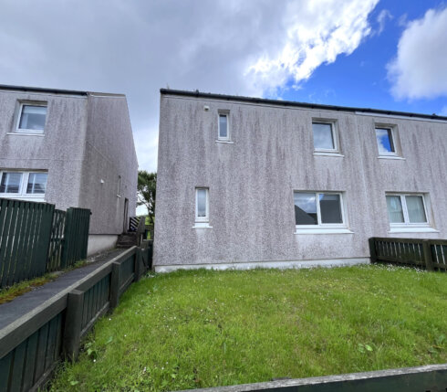 Two adjacent white residential buildings with small windows, separated by a narrow pathway and green lawns. A wooden fence borders the properties. The sky is partly cloudy.