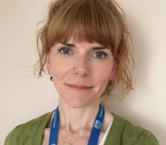 A person with auburn hair and a fringe is wearing a green cardigan and a blue lanyard. They are standing in front of a beige background.