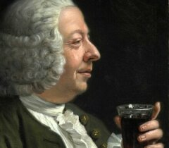 A man with white curly hair and formal clothing holds a glass of dark liquid, looking at it with a slight smile against a dark background.