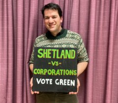 A person stands in front of a purple curtain holding a sign that reads, "SHETLAND vs CORPORATIONS VOTE GREEN.
