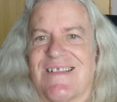 Close-up portrait of a smiling person with long, wavy silver-grey hair, showing a gap between their front teeth, against a light wooden